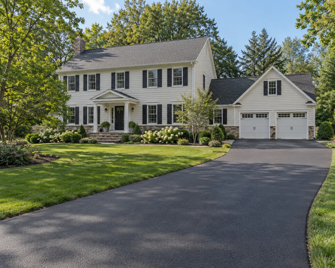 Ivory colonial home with a freshly paved asphalt driveway, landscaped lawn, and mature trees